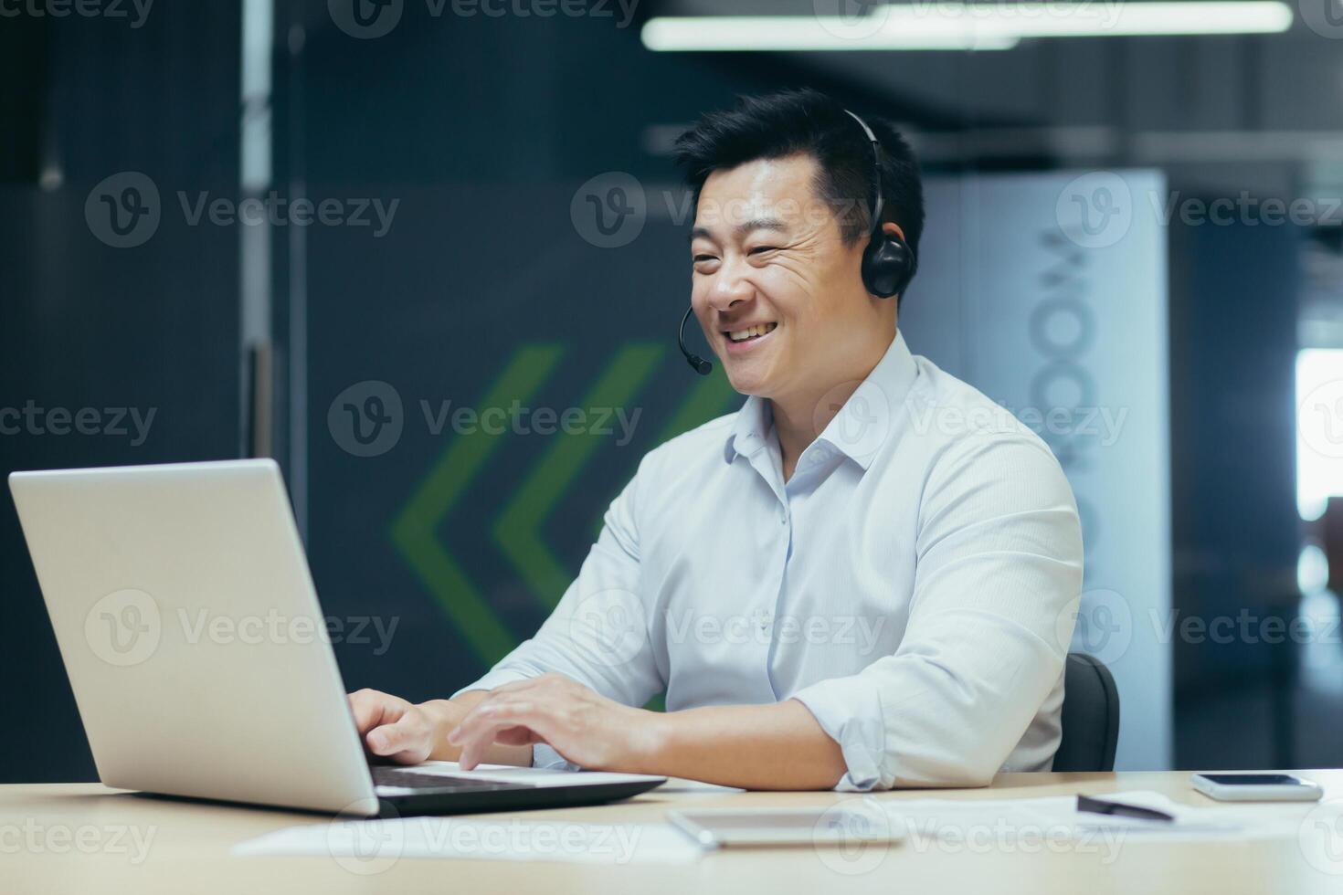 Portrait of a smiling young Asian man working in the office at a laptop in a headset. photo