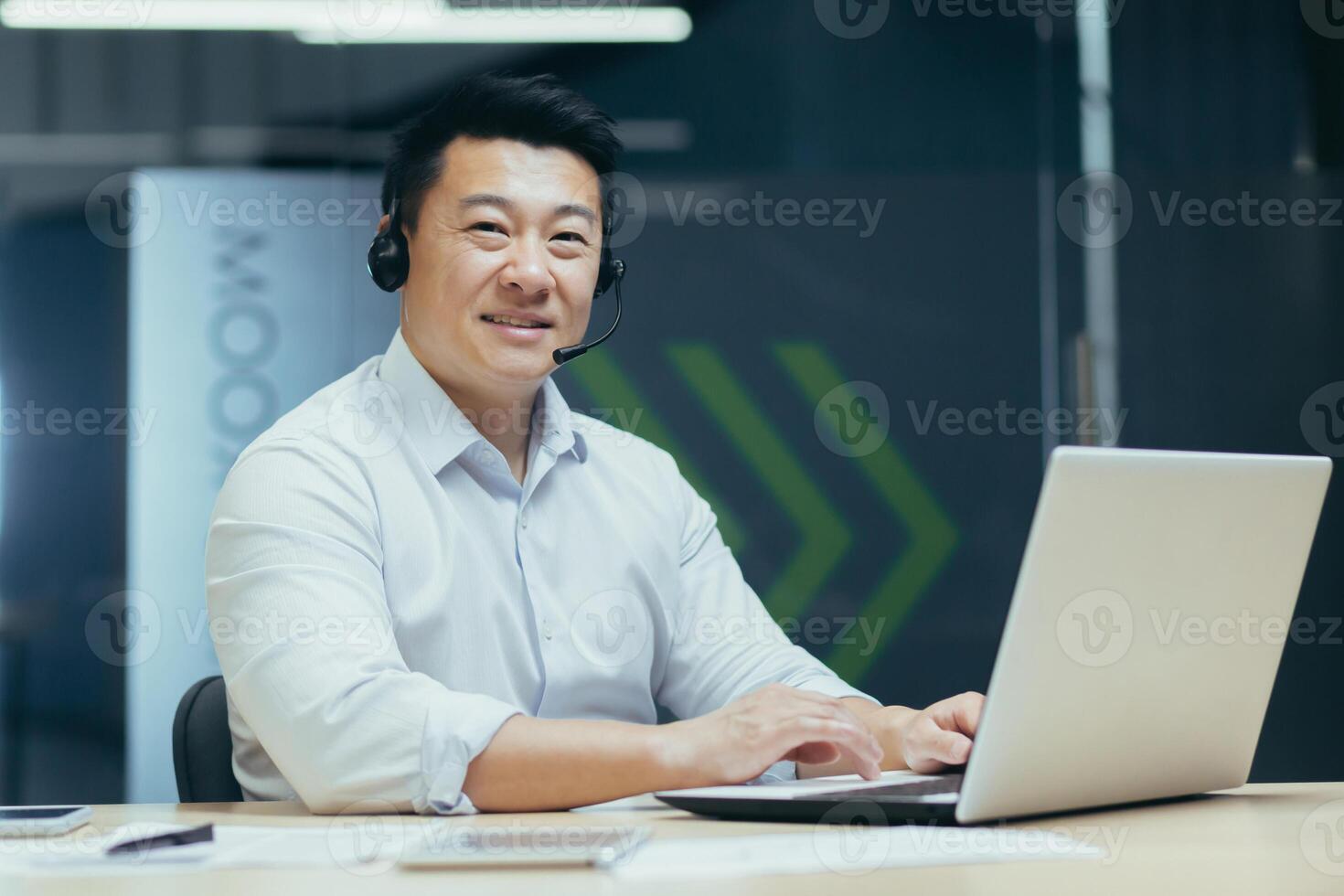 Portrait of successful asian businessman man with call headset looking at camera and smiling, tech support consultant working in office with laptop photo