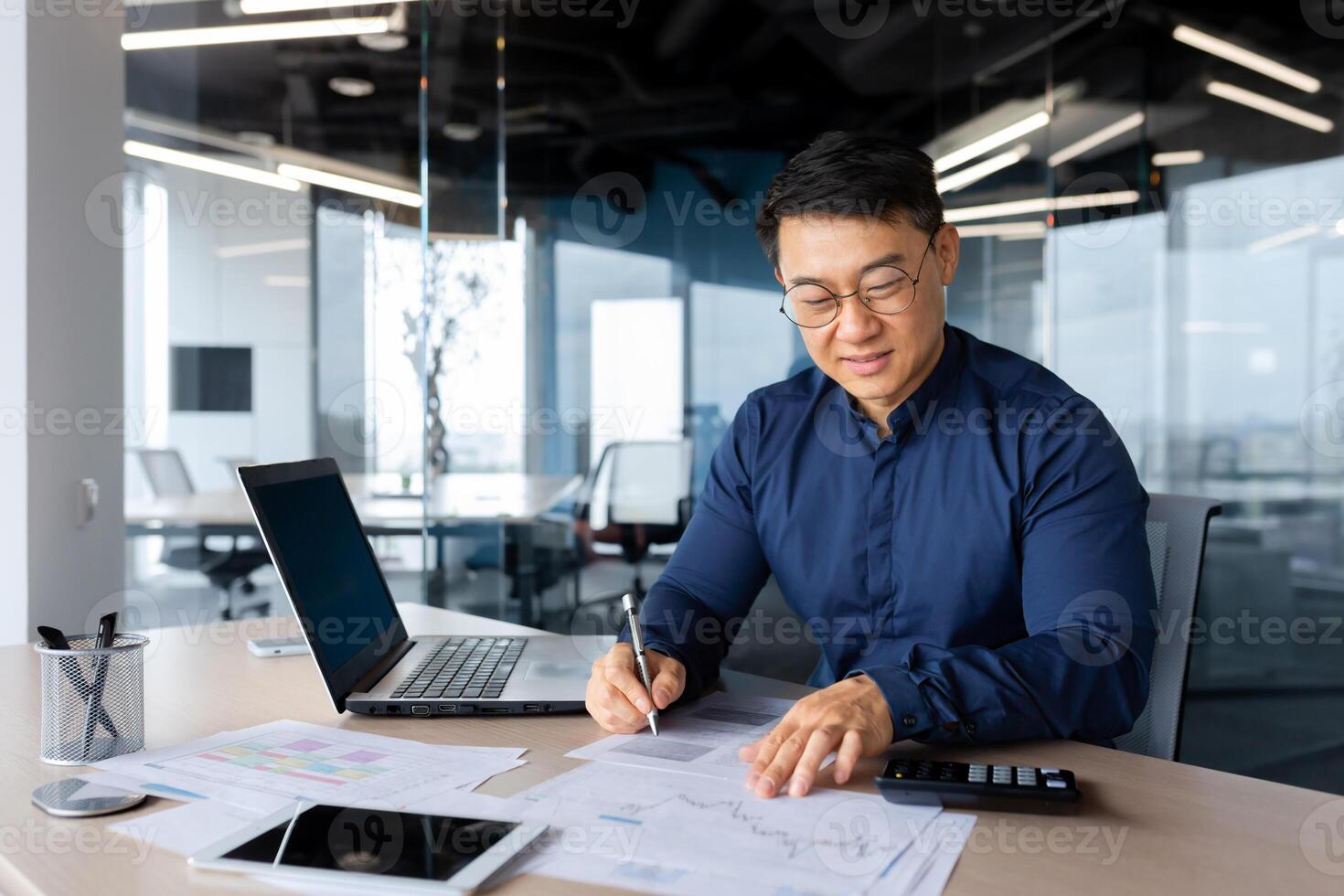 Successful and happy investor working with documents and charts inside office, Asian businessman behind paper work calculates bills with calculator, man at work with laptop happy with achievements. photo