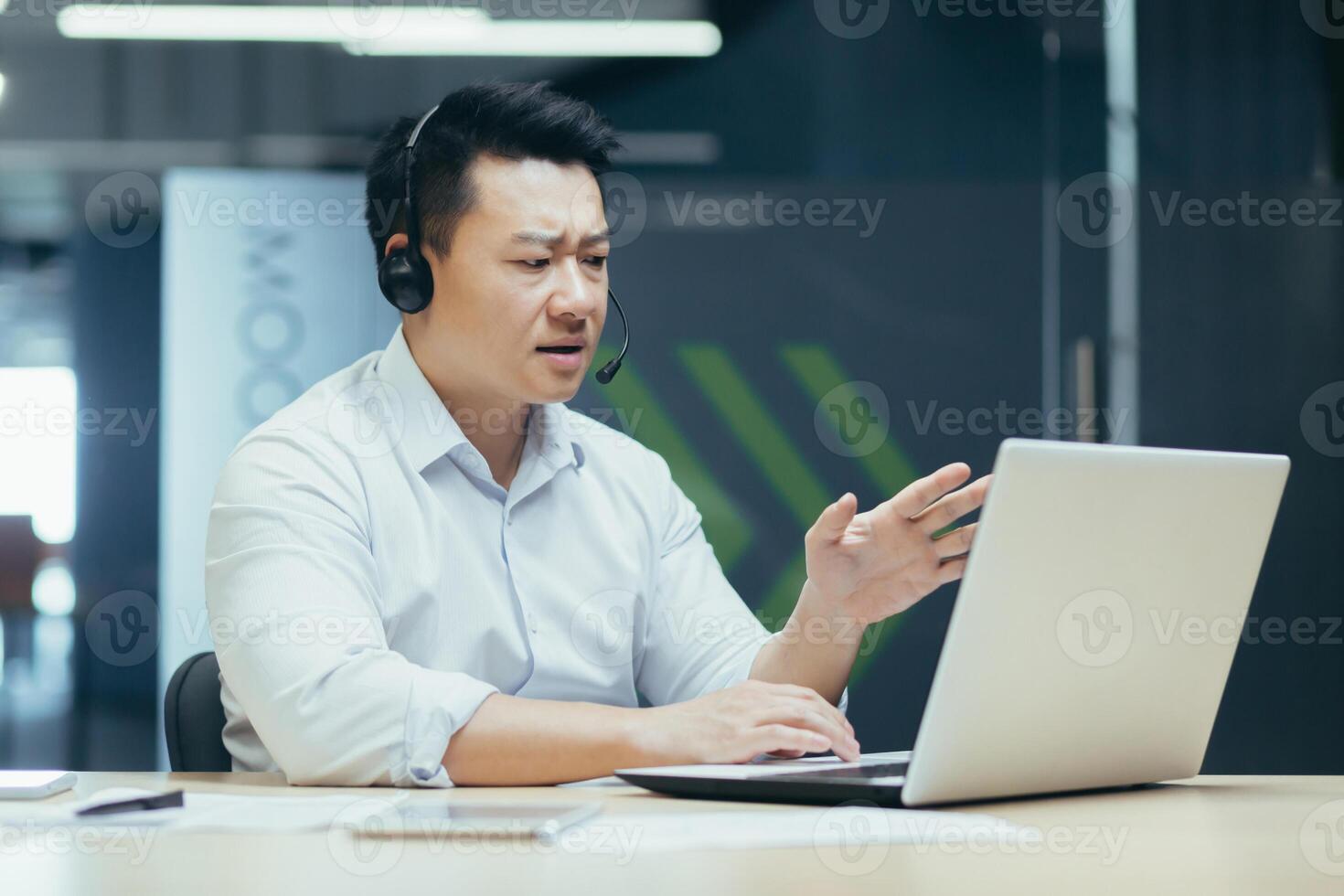 Young Asian man, sales consultant, support service agent in headset. Sits in the office at the laptop and consults, talks through a call from the laptop. photo