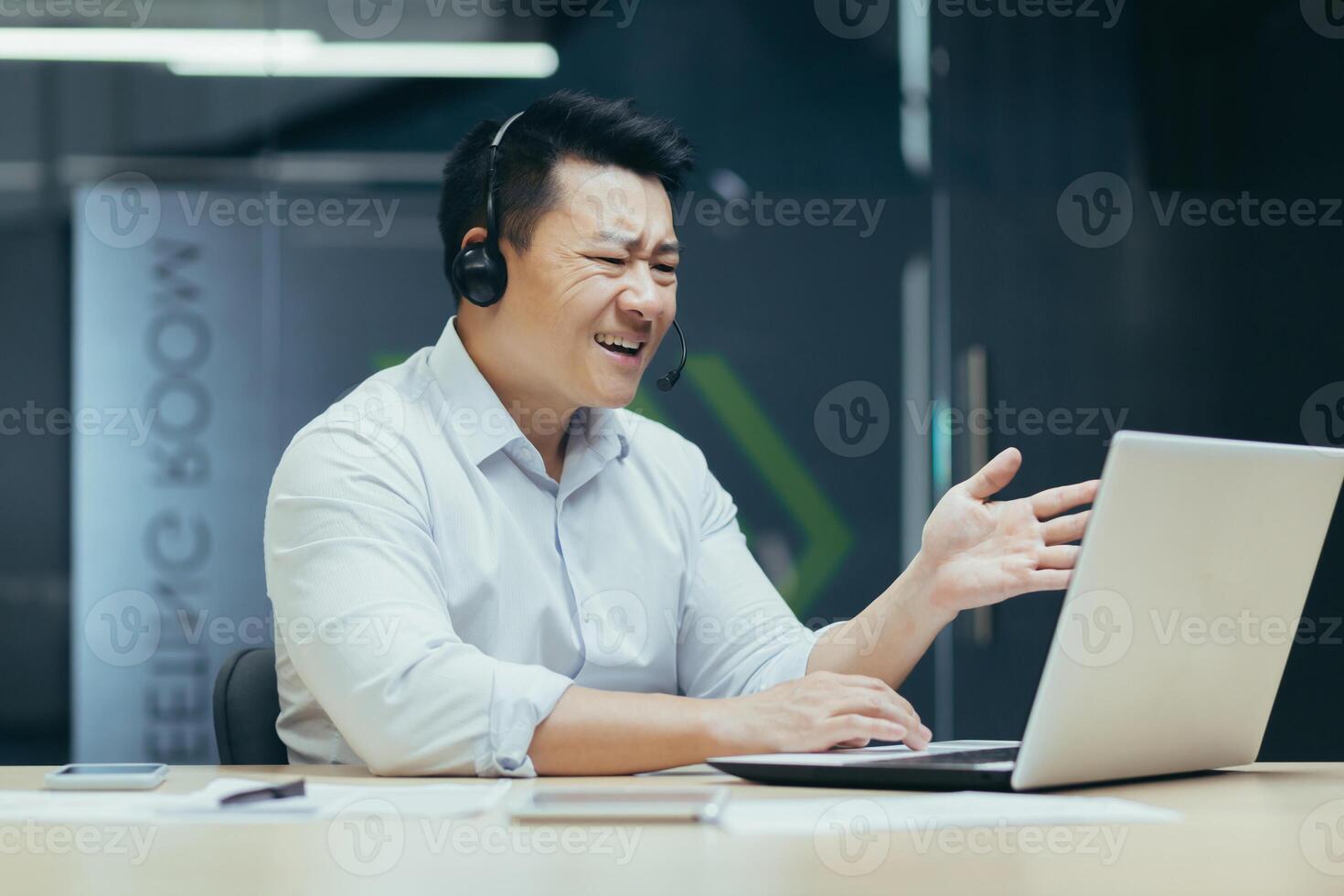 Angry and annoyed asian businessman with headset for call, talking in online meeting, looking at laptop screen, man at work photo