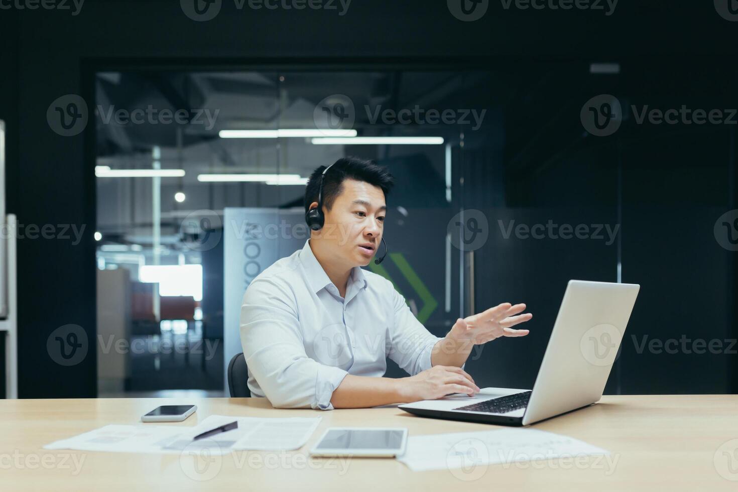 Portrait of a young Asian man in a headset. Sits in the office at the laptop and consults, talks through a call from the laptop. photo