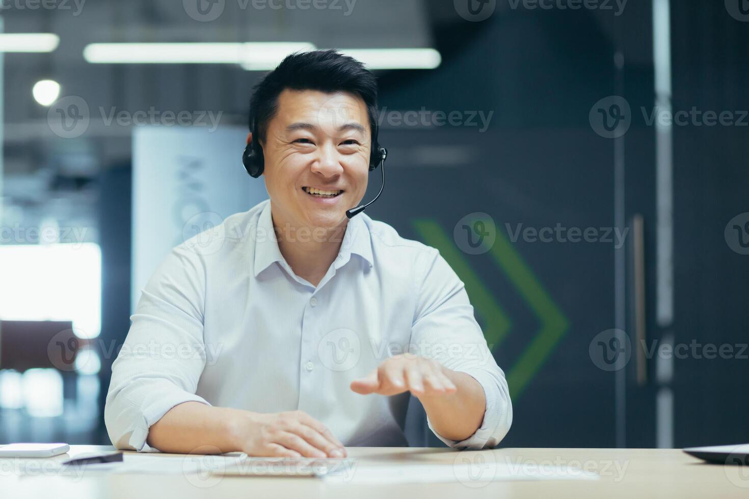 Online business training. Young smiling Asian man sit and talk in the office in front of the camera in a headset, conducts an online meeting, webinar photo