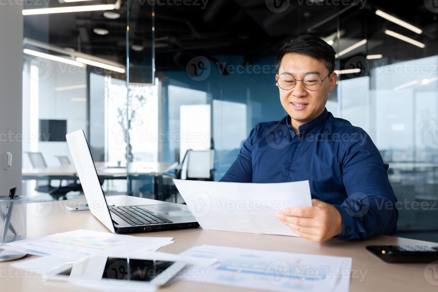 A young man, an Asian architect, designer, engineer, freelancer works in the office at a desk using a laptop. Holds documents, plans, drawings in his hands. photo