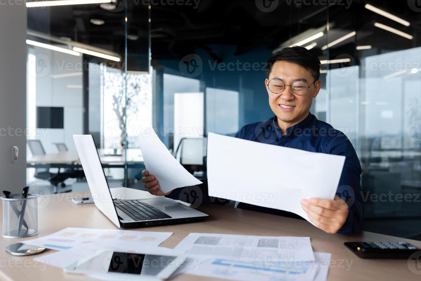 A young Asian architect, designer, engineer. Sitting at the desk in the office, holding papers. Works with projects, plans. Studying documents, deal. photo