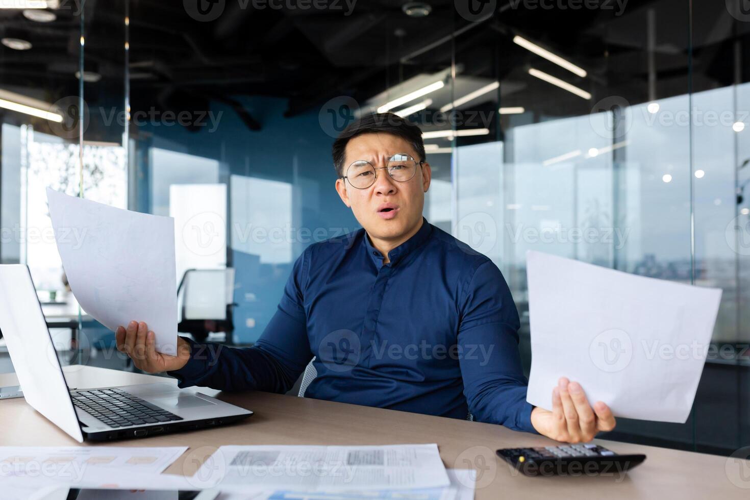 Problems at work. Worried young Asian man, businessman, freelancer. He sits in the office at a table with a laptop, holds documents in his hands. Spreads his hands, looks at the camera confused. photo
