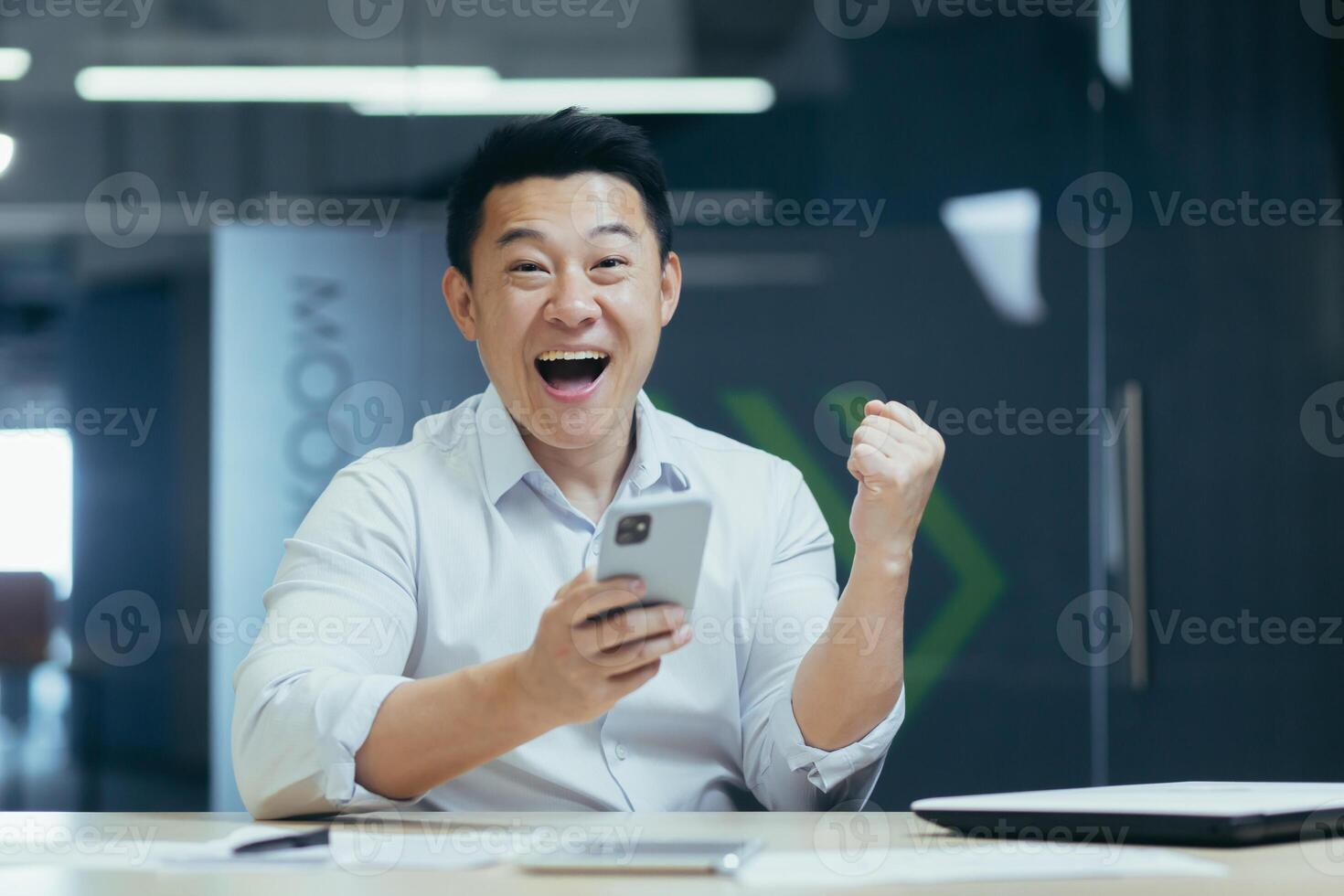 A young man, an Asian office worker, plays online games on the phone, rejoices at the victory. He points with his hand, yes, the winner. Sitting in the office at the desk, looking at the camera. photo