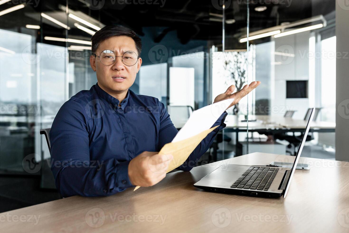 Annoyed asian businessman looking at camera and showing received letter with bad news, man working inside modern office building, using laptop. photo