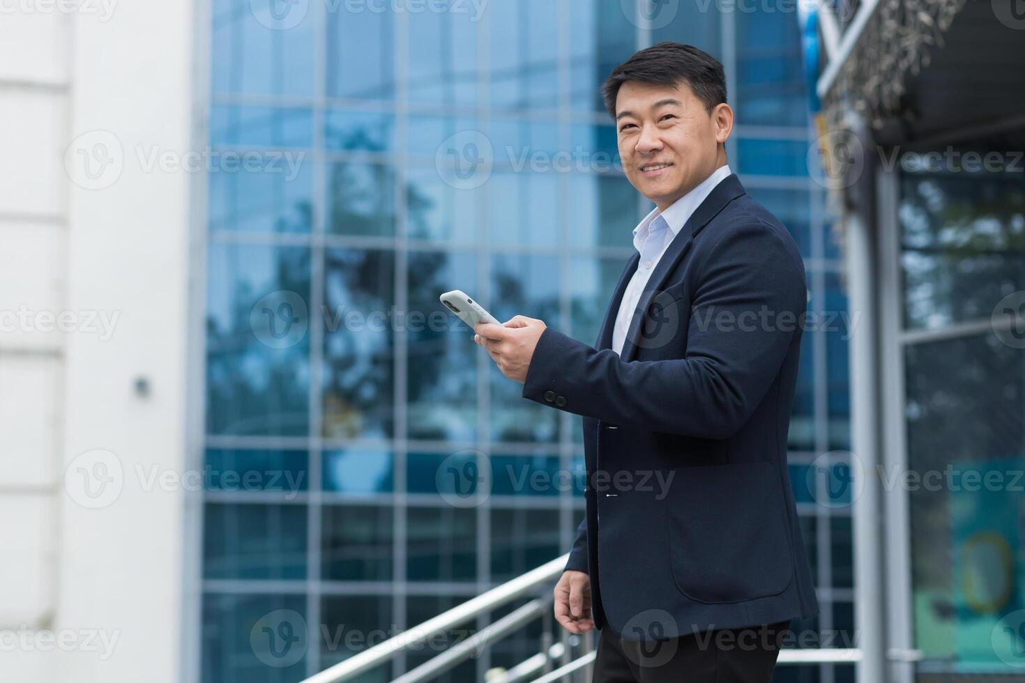 Portrait of an Asian businessman outside an office building, a man in a business suit is using a smartphone, smiling and looking at the camera, walking down the street. photo