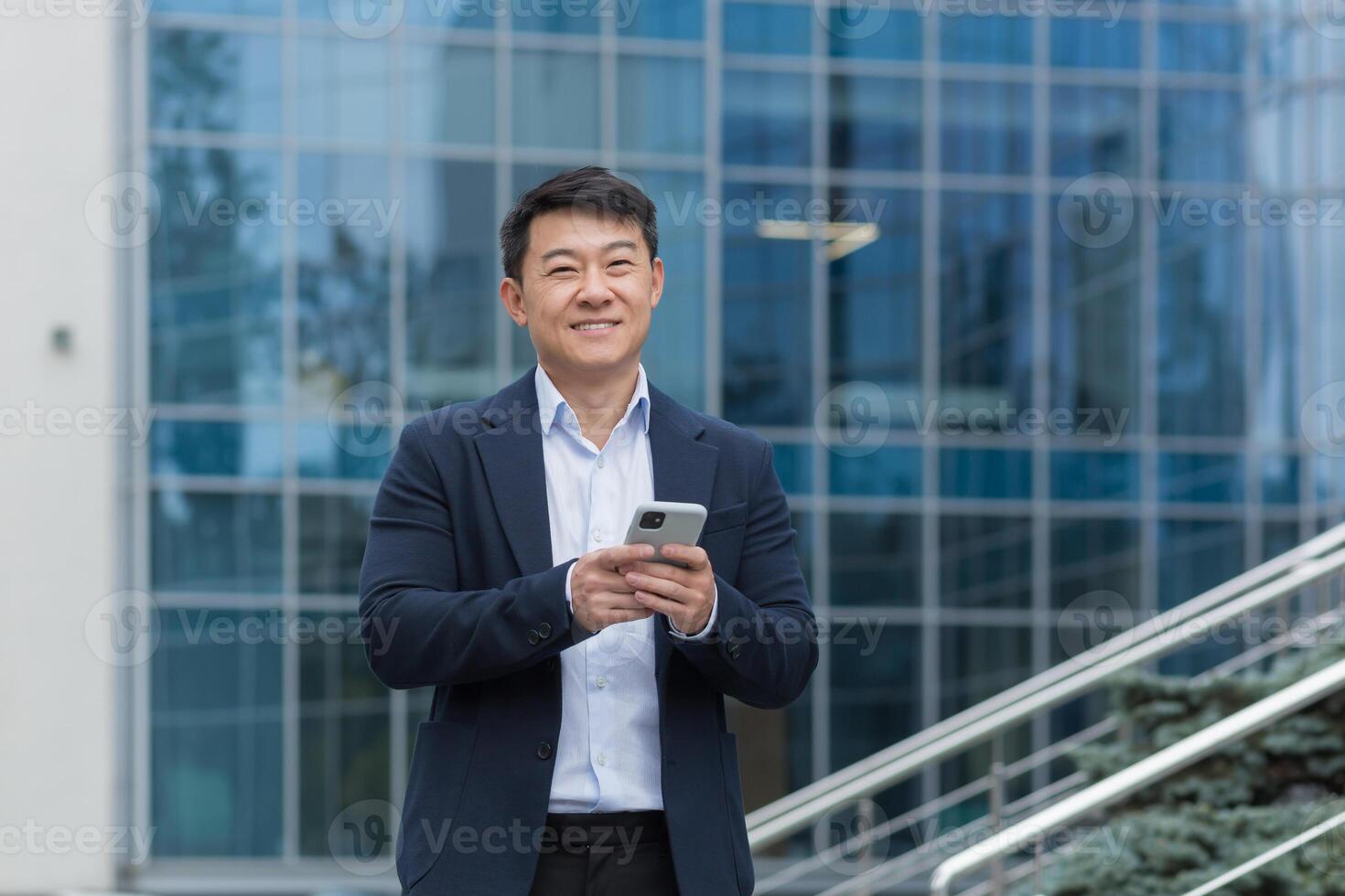 Portrait of Asian businessman outside modern office building, boss in business suit using phone smiling and looking at camera, man typing message and browsing web pages online. photo