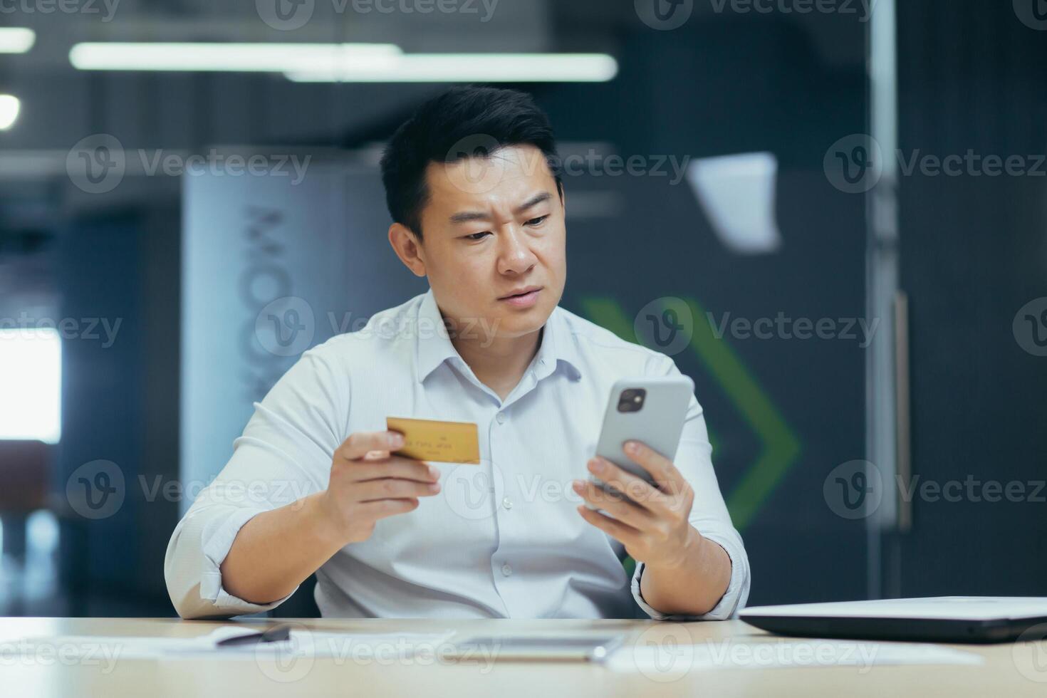 Worried young asian man sitting at desk in office and holding phone and blocked credit card, financial problems, unsuccessful shopping, money transfer. photo