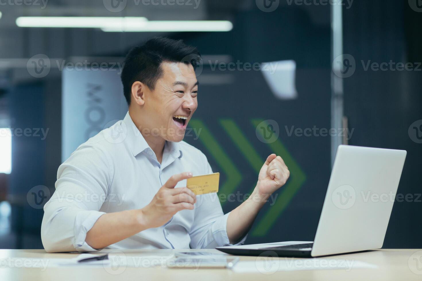 Happy young handsome man Asian businessman rejoices, received income,salary on credit card. He works with a laptop, holds a credit card, sits at a desk in the office photo