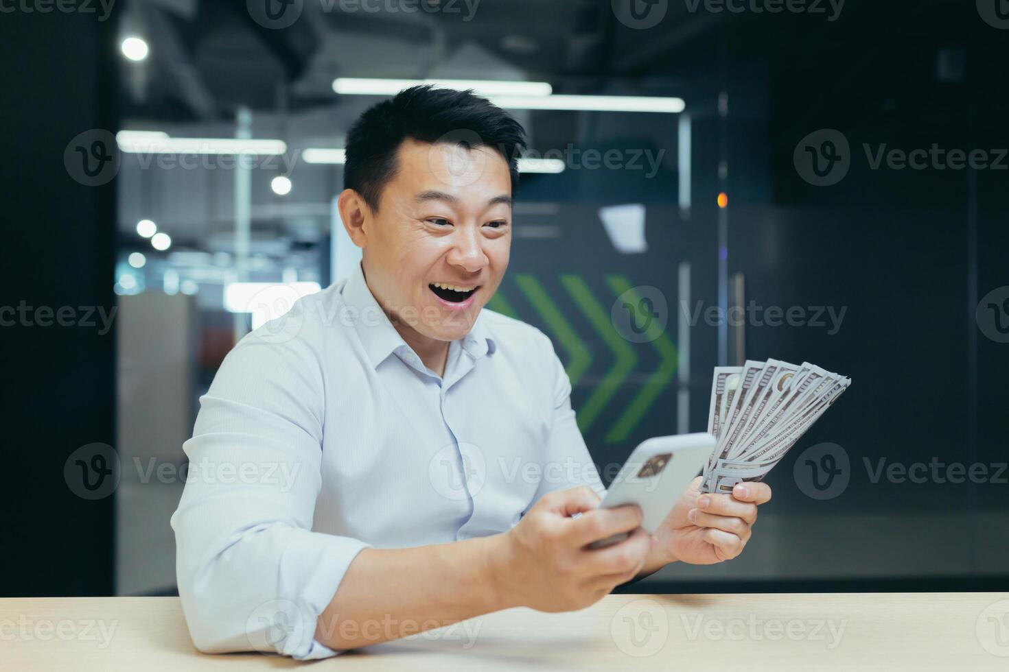 Young smiling man, Asian financial businessman sitting at the desk in the office and using the phone. He is holding a fan of banknotes in his hands, looking at his mobile phone in surprise. photo