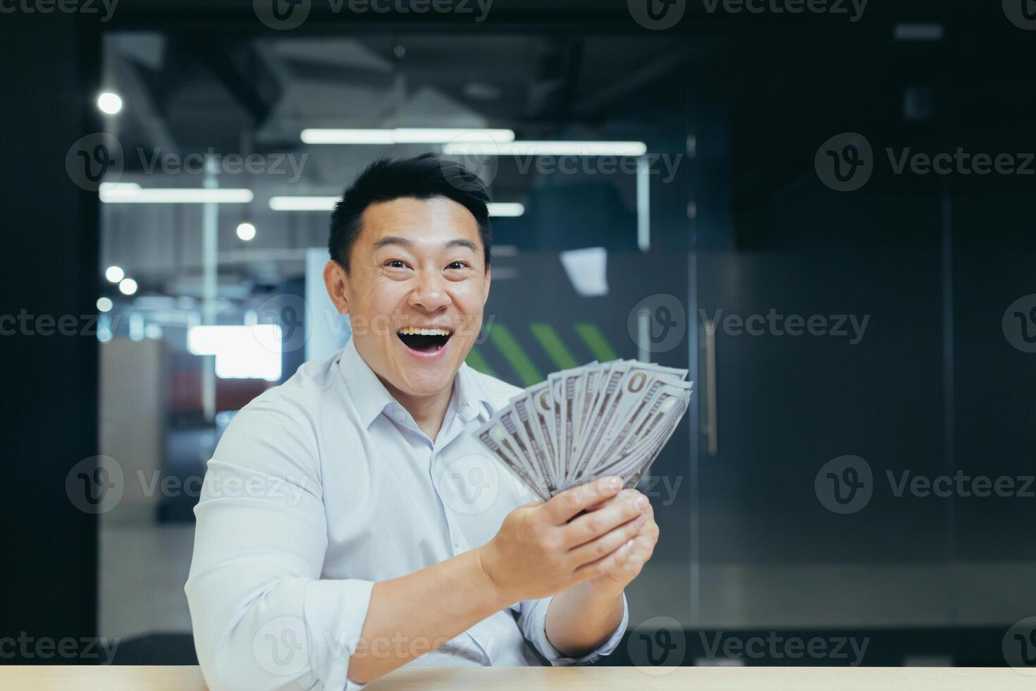Shocked young Asian man holds a lot of money in his hands, cash, dollars. He looks surprised and happy at the camera, sits at the table in the office. photo