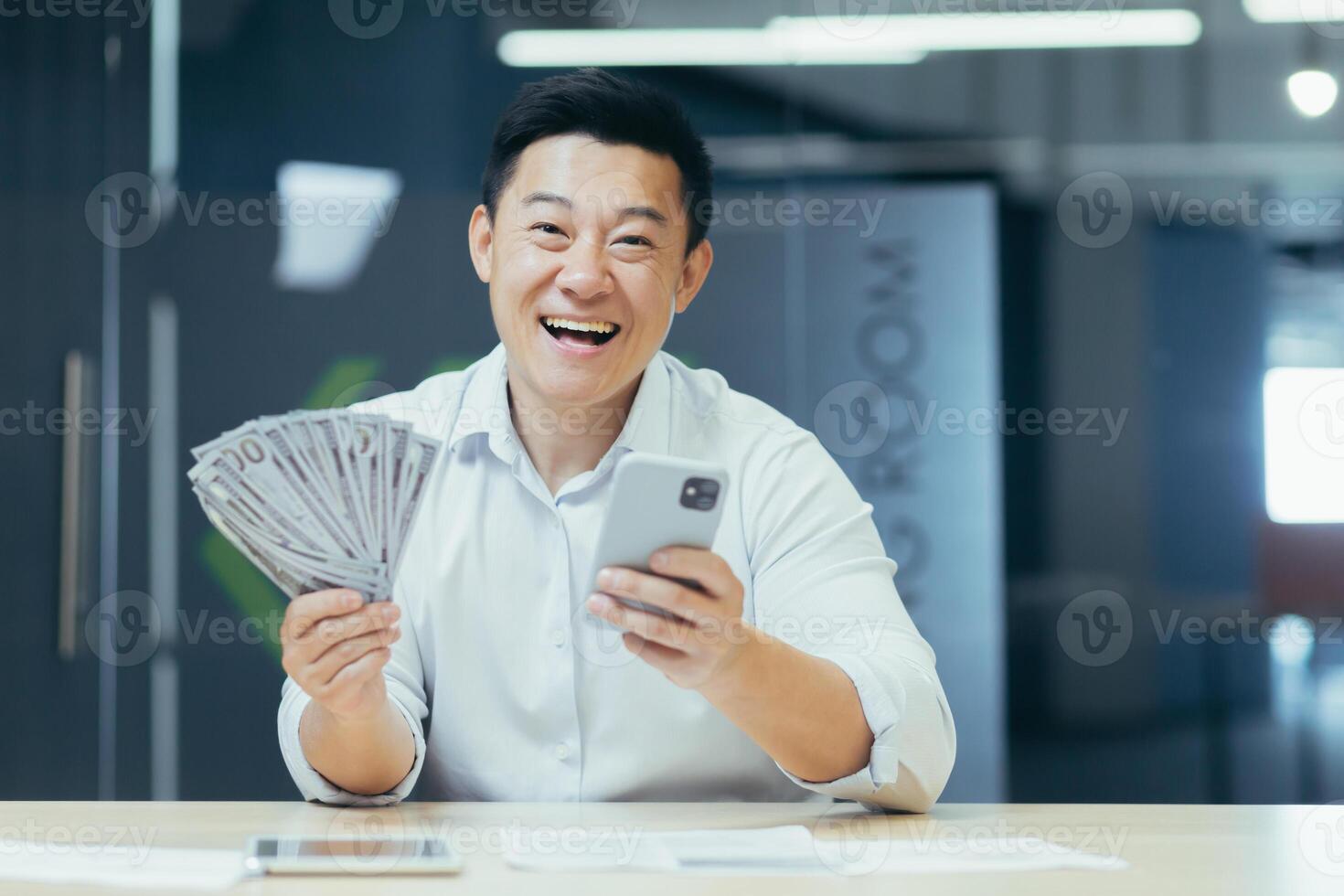 A happy young Asian man sits in the office at the table, holds a phone and cash money in his hand. A smiling man dials the phone, rejoices at a monetary win, lottery. photo