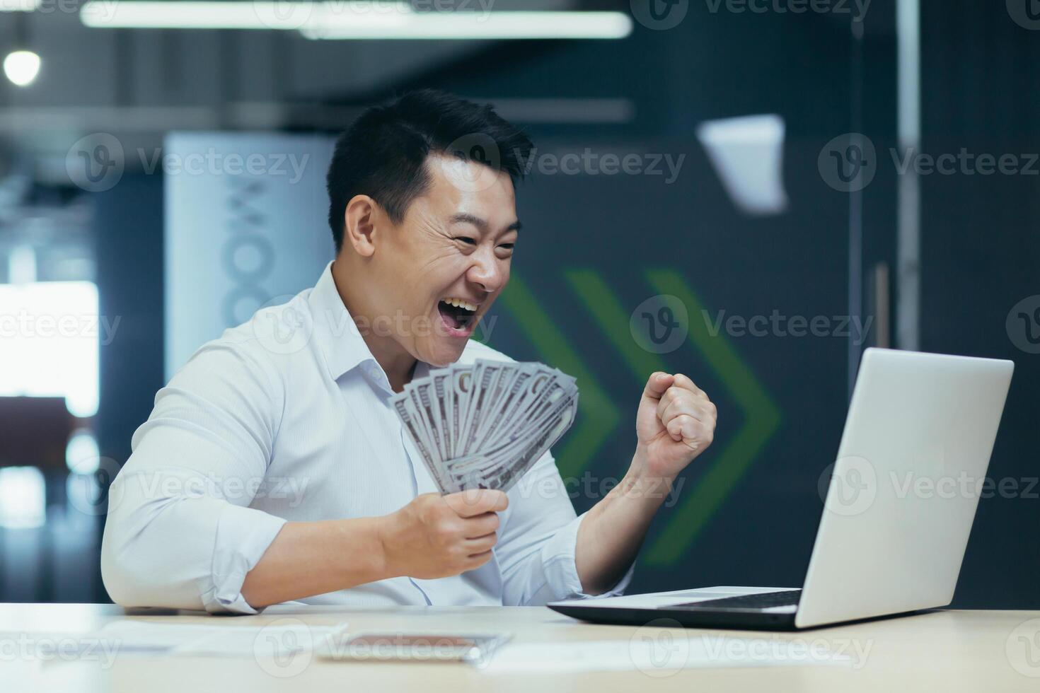Successful Asian investor businessman, happy with victory and conclusion of investment contract, man holds dollars cash in hand and shouts joyfully at laptop screen photo