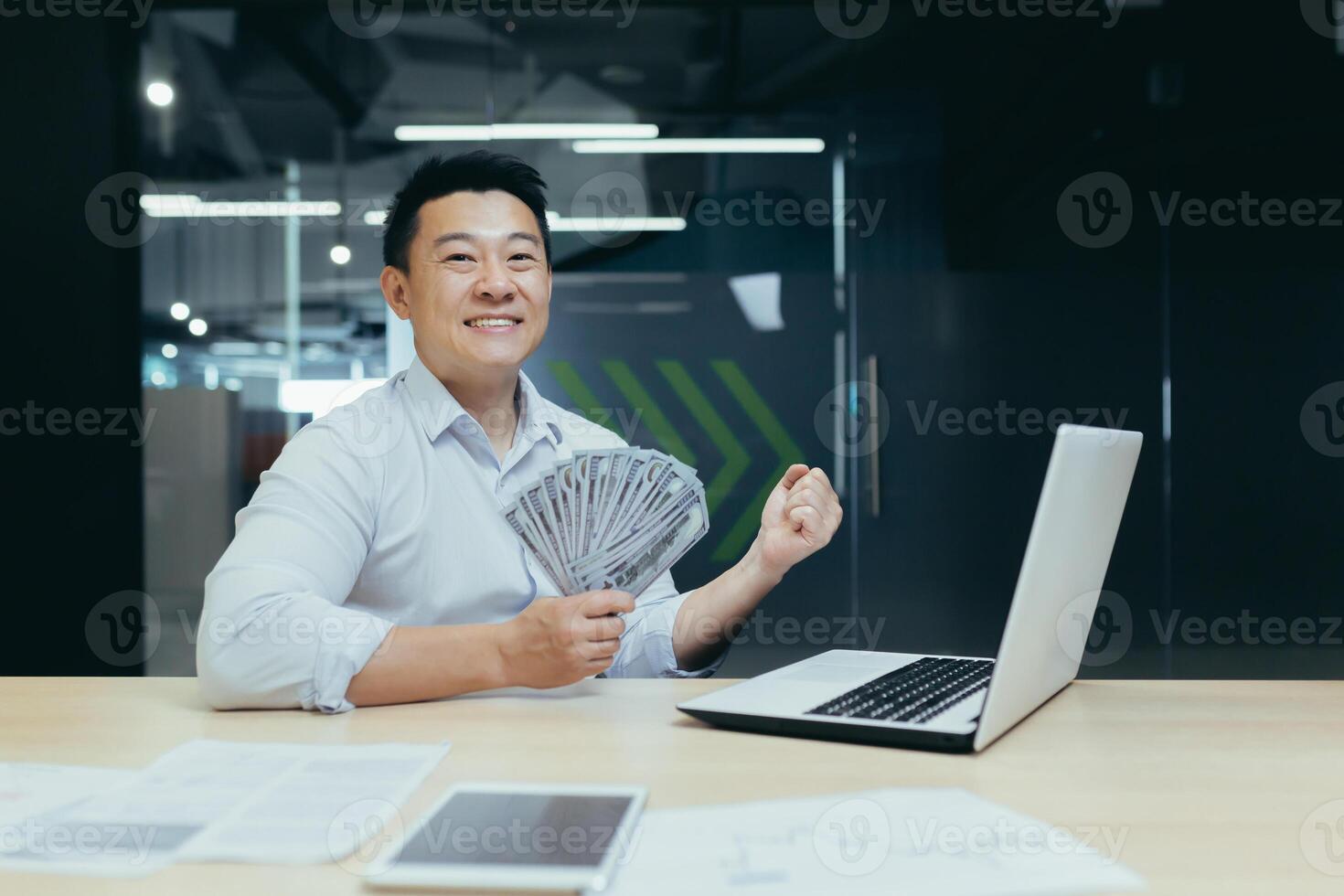 Portrait of a happy Asian man working in the office on a laptop, holding a fan of cash, dollar bills in his hand. shows a victory gesture with his hand to the camera, celebrates. photo