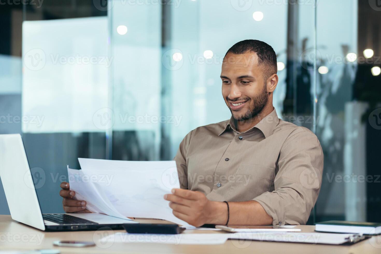 Successful businessman inside the office reading a financial report, hispanic man holding a contract in his hands and smiling with satisfaction, good achievement results, working on a laptop. photo