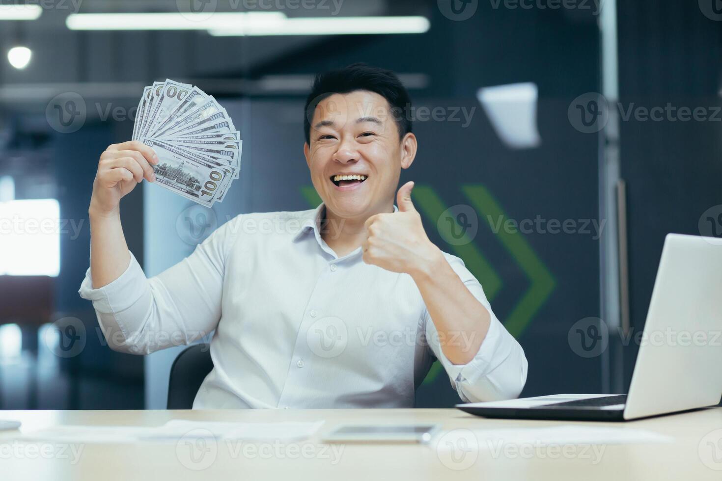 A happy young Asian man sits in the office at a table with a laptop, holds a fan of money, cash in his hands, shows a super finger. The smiling man looks and points to the camera. photo