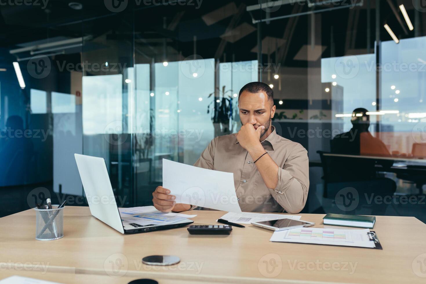 Thoughtful young African-American male designer, architect sitting in the office at the table with a laptop. He holds documents in his hands, concentrates on working with plans and drawings. photo