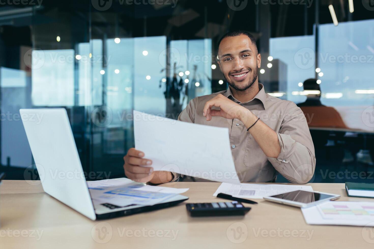 A young African-American male designer, architect sitting in the office at the table with a laptop. He holds documents in his hands, working with plans and drawings, looks at the camera, smilling photo