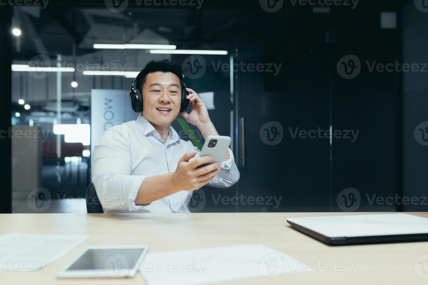 A young Asian man in headphones sits at the office table, holds the phone in his hands, looks at the screen, selects a playlist, a podcast. listening to music, relaxed, break and rest at work, study. photo