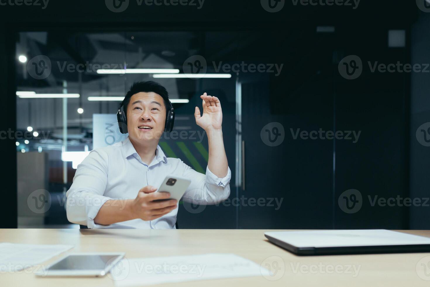 Rest, break at work. A young Asian man in headphones sits at the office table, holds the phone in his hands and listens to music, dances, relaxed. photo