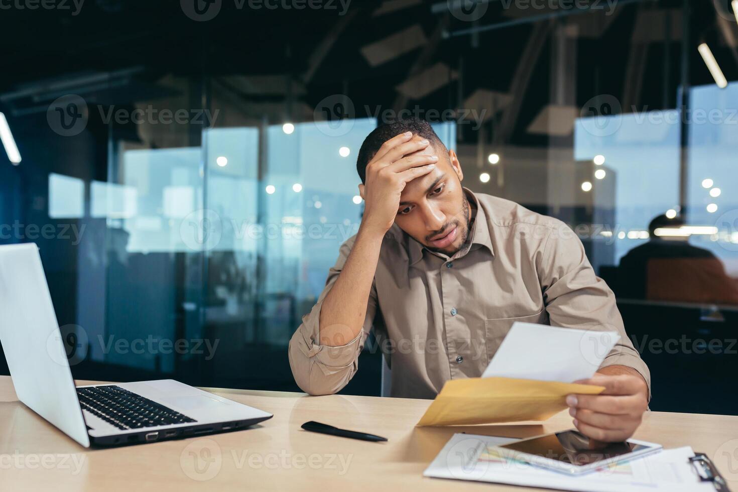 Pensive businessman inside office reading notification letter, man received envelope thinking about decision sitting at table and laptop inside office, bad news message. photo