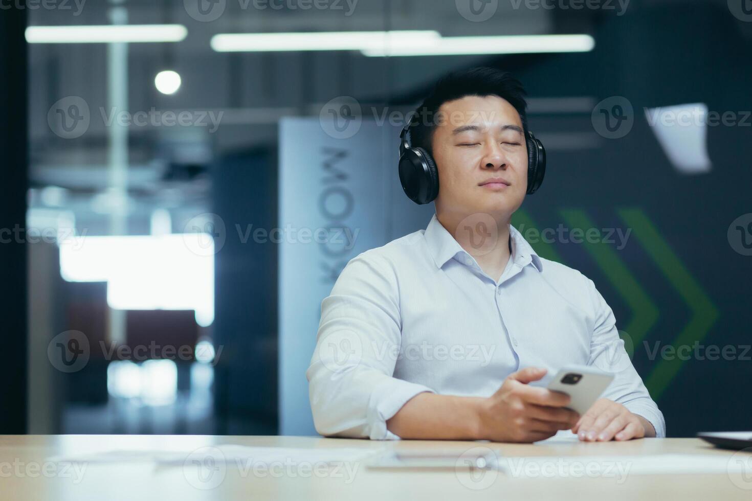 Asian boss business owner relaxing in office, listening relaxing and relaxing music, man sitting at desk, using big headphones and online listening app on phone photo