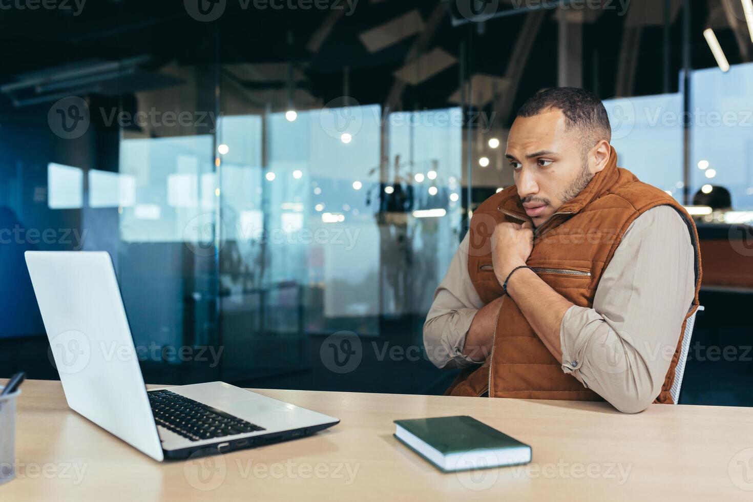 African american man freezing in office, no heating in workplace, man in outerwear sitting at desk using laptop at work. photo