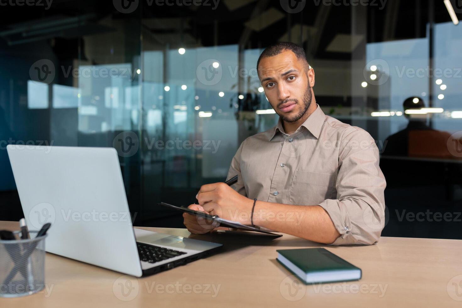 Portrait of serious and focused businessman, man looking thinking at camera writing and signing document inside office, african american financier at work on laptop. photo