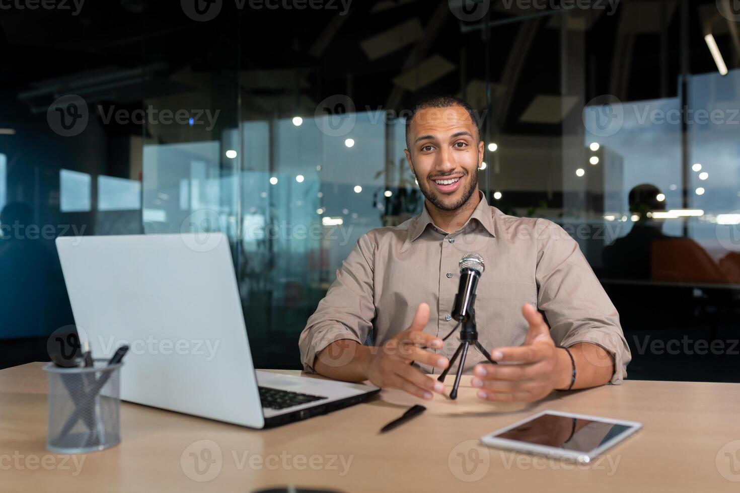 Portrait of a successful African American influencer, man smiling and looking at the camera, using a laptop and a professional microphone at work to record podcasts and read audio books online. photo