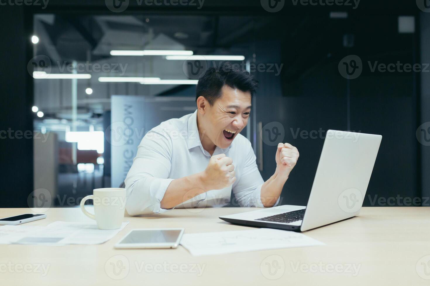 Portrait of successful Asian boss, businessman looking at laptop screen reading good news and celebrating victory and success, man at work photo