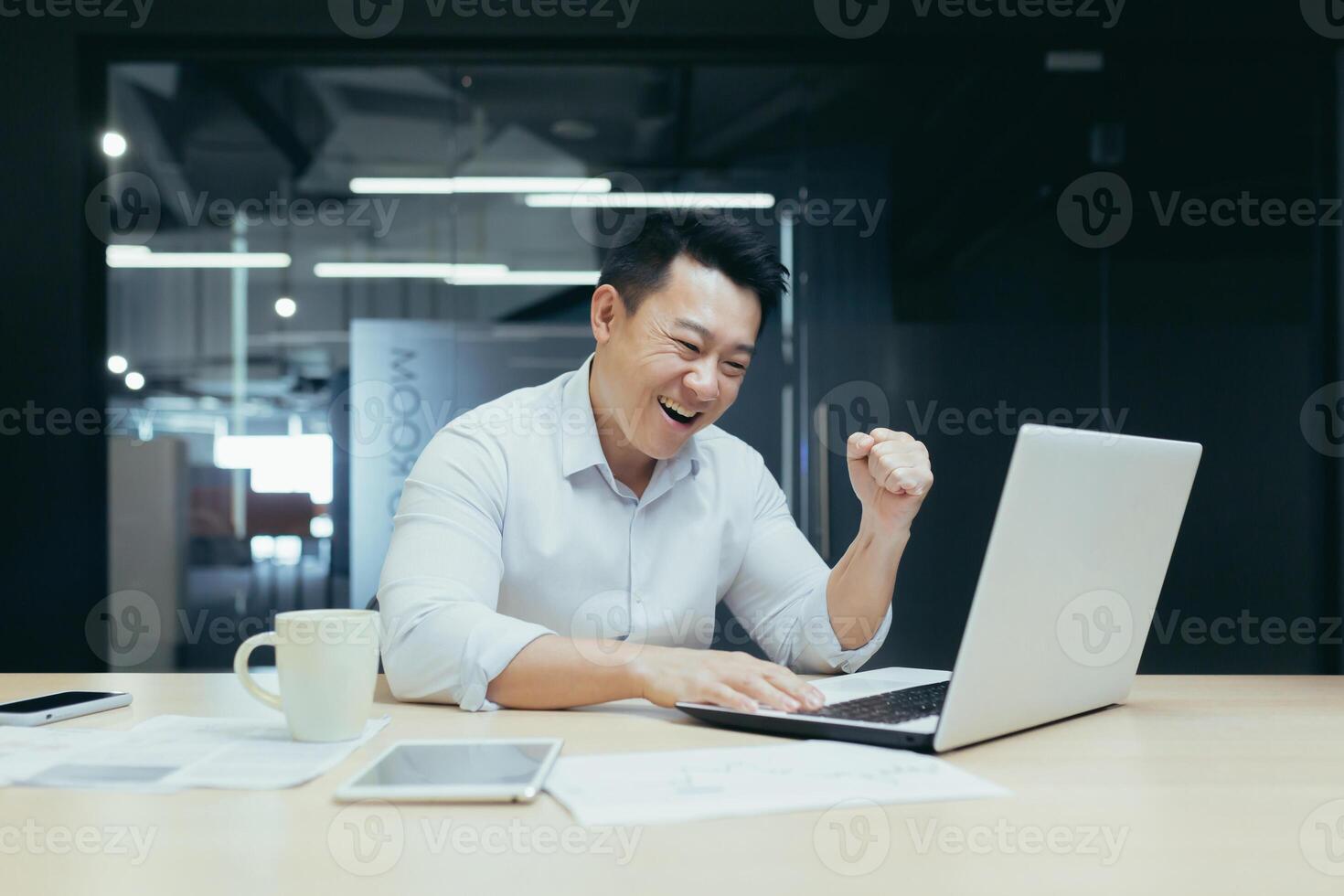 A young Asian man sits in the office at the table, rejoices at the victory and the win, looks at the laptop, participates in the auction, places bets. He smiles, shows a yes gesture with his hand photo
