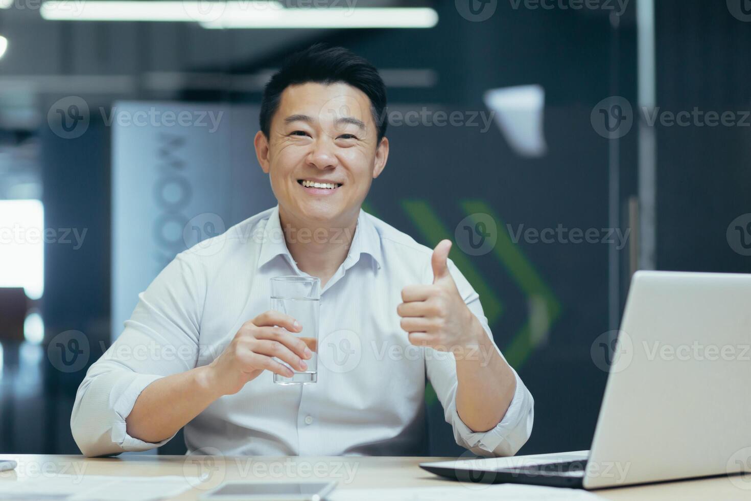 A young Asian freelancer man drinks water in the office at his desk, maintains water balance. He holds a glass in his hands, looks at the camera with a smile, and points a super finger. photo