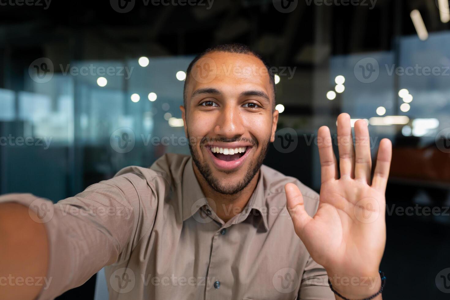 Smiling and cheerful businessman in office taking selfie photo on phone and talking on call with colleagues and friends using smartphone, african american man waving at camera greeting gesture.