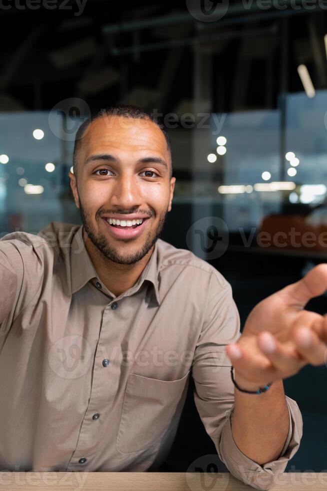 ringing, successful businessman looking at smartphone camera talking remotely with colleagues, Hispanic man smiling at work inside office, webcam view pov vertikal photo