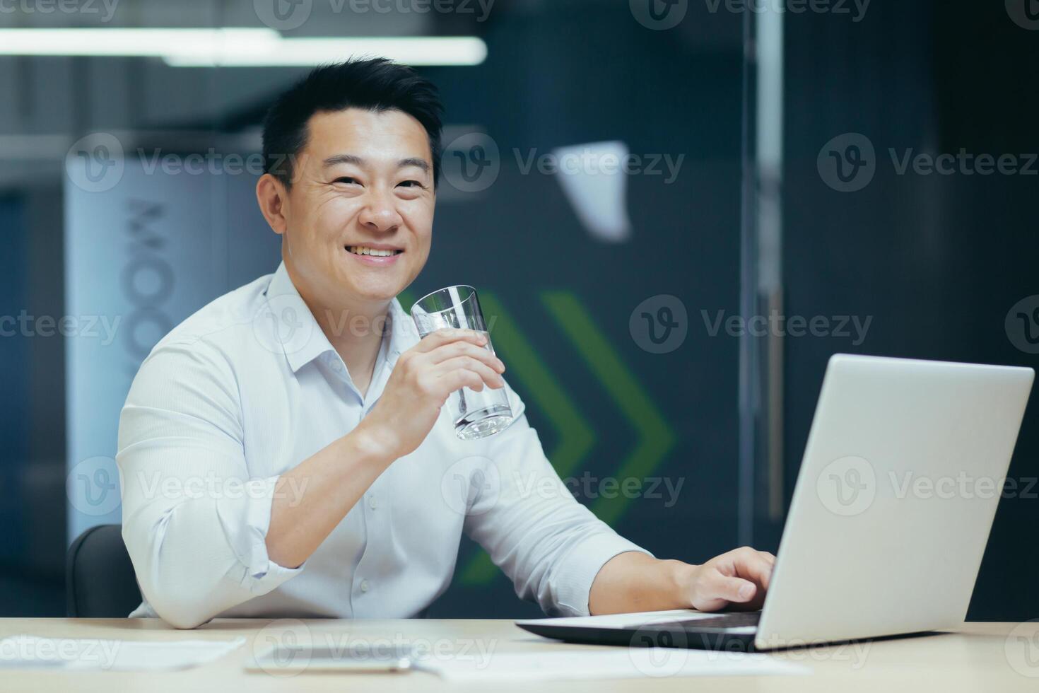 Asian businessman in the office drinking clean filtered water, portrait of a man smiling and looking at the camera holding a glass of water, a man at work with a laptop photo