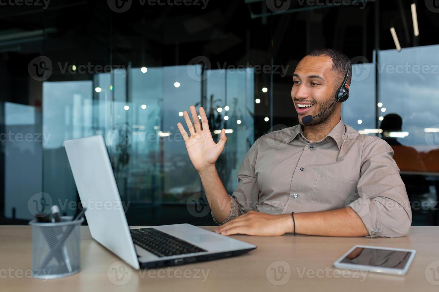 Online training. A young African-American male student sits in an office at a desk in front of a laptop in a headset. Learns through a call, says hello, smiles. photo
