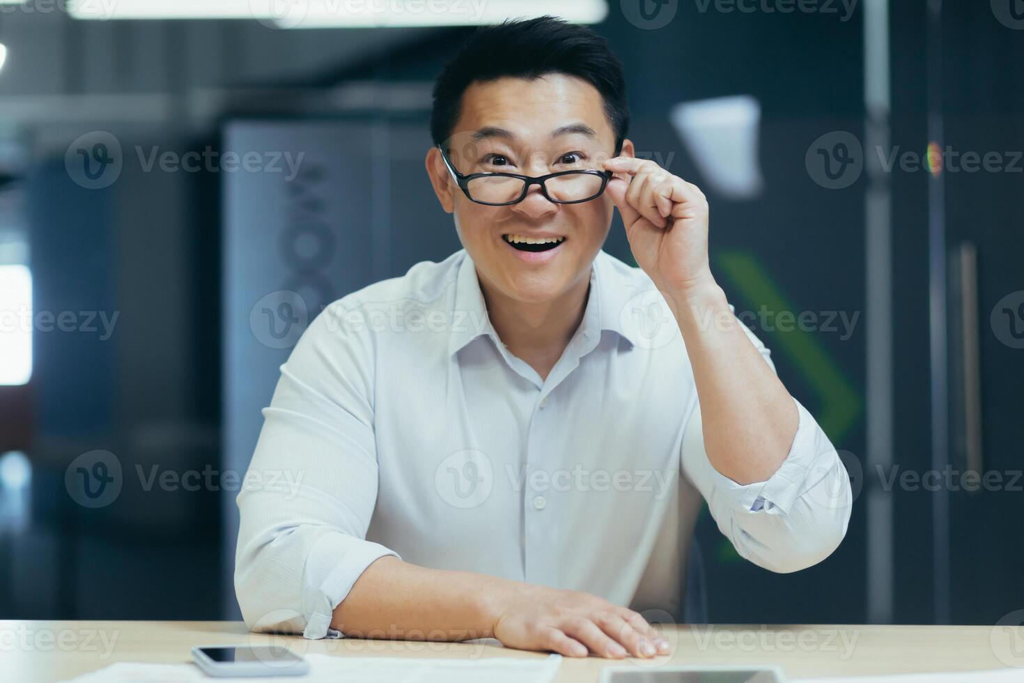 Portrait of a happy young Asian man sitting in the office at the table with phone and documents, holding glasses in his hand, looking down, smiling, looking at the camera. photo