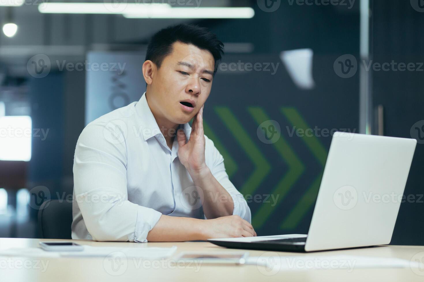Toothache at work. A young Asian man is sitting at the desk in the office and holding his cheek. He feels a strong toothache, discomfort, grimaces. photo
