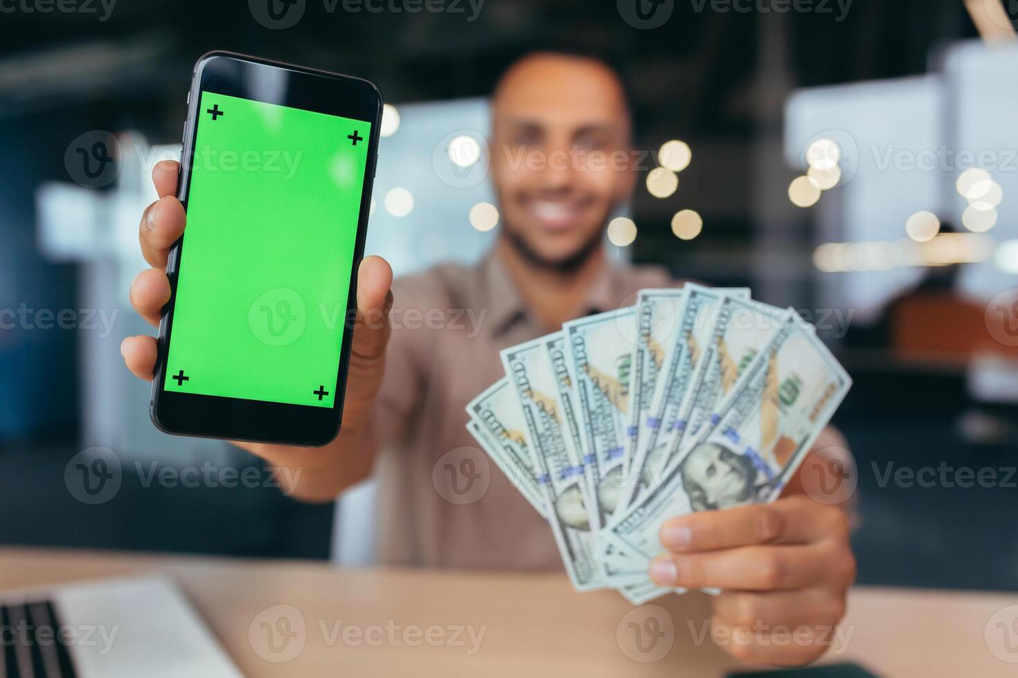 Selective focus, businessman working inside modern office building, african american man with laptop looking at camera showing smarphone ekras and winning dollars money cash. photo