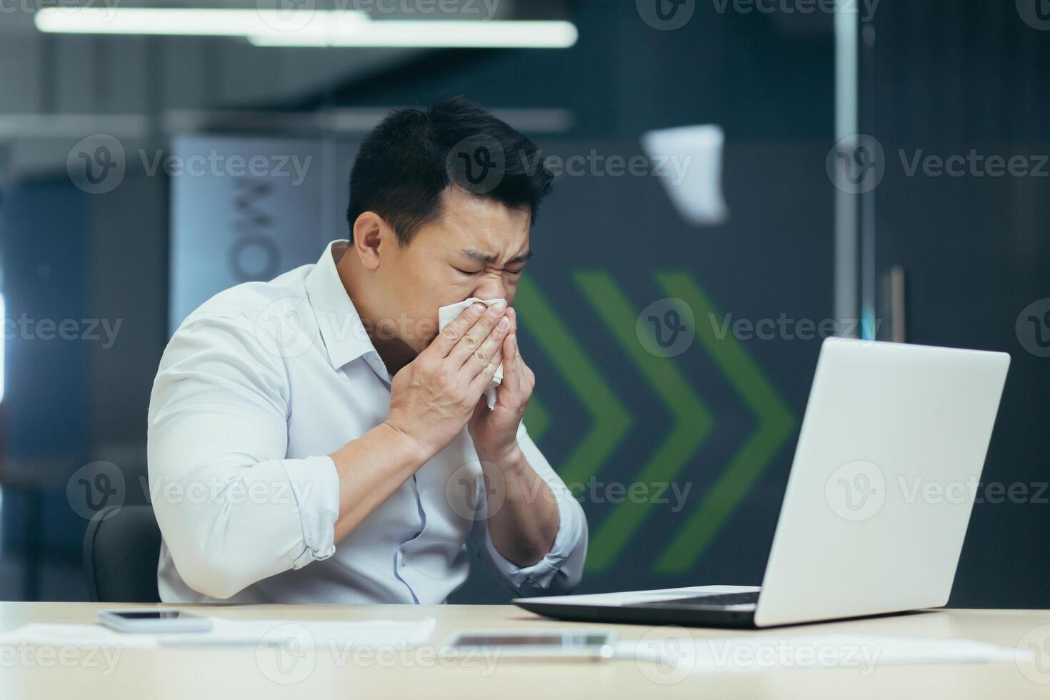A young sick Asian man is sitting at work in the office. He wipes his nose from a runny nose with a napkin, coughs, feels bad. photo