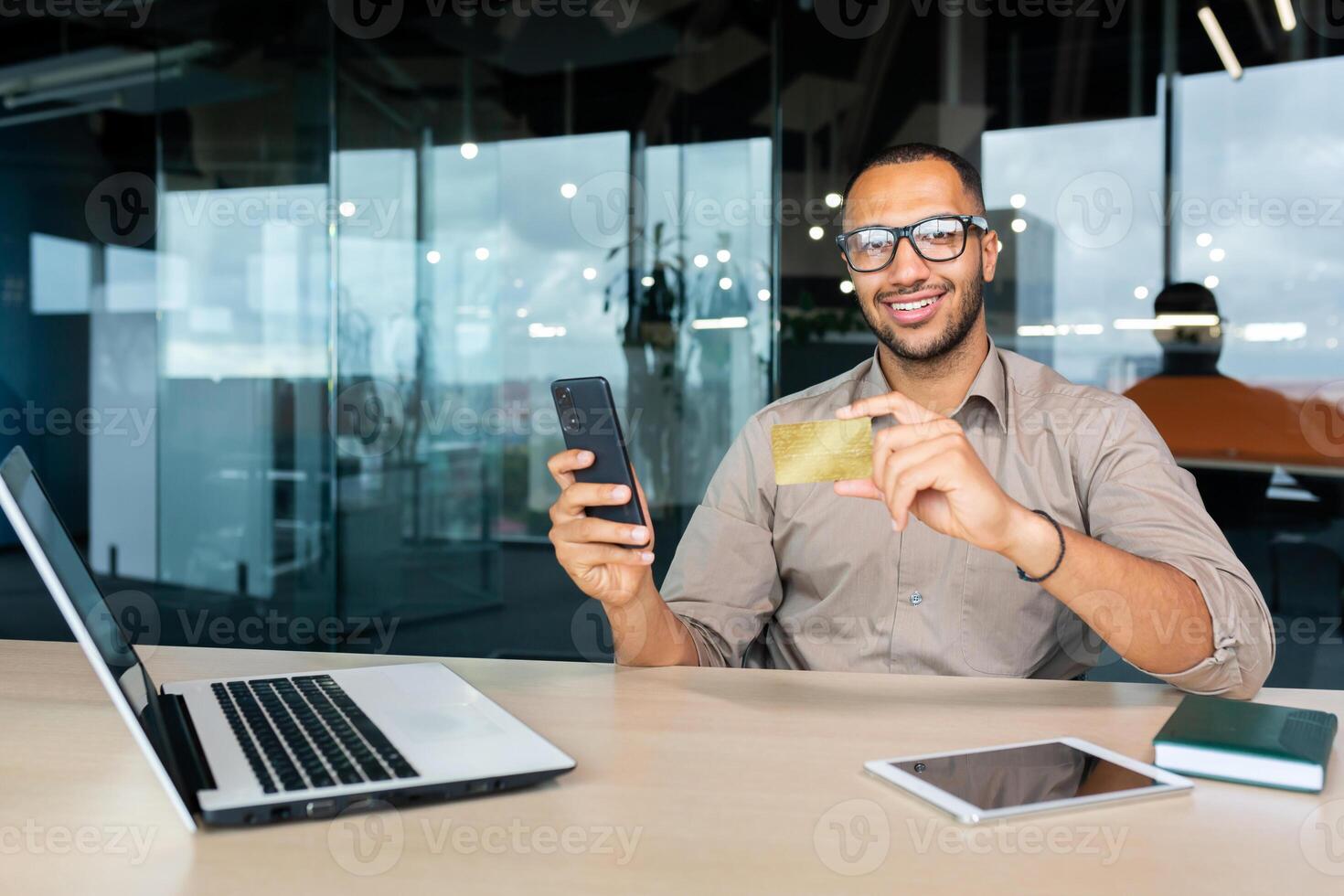 Portrait of successful African American businessman inside office, man smiling and looking at camera, worker holding phone and bank credit card working at work using laptop. photo