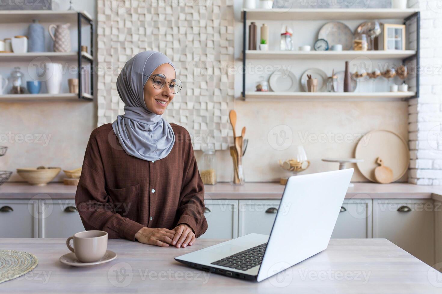 Young beautiful muslim woman in hijab and glasses studying remotely using laptop student woman in kitchen smiling and happy. photo