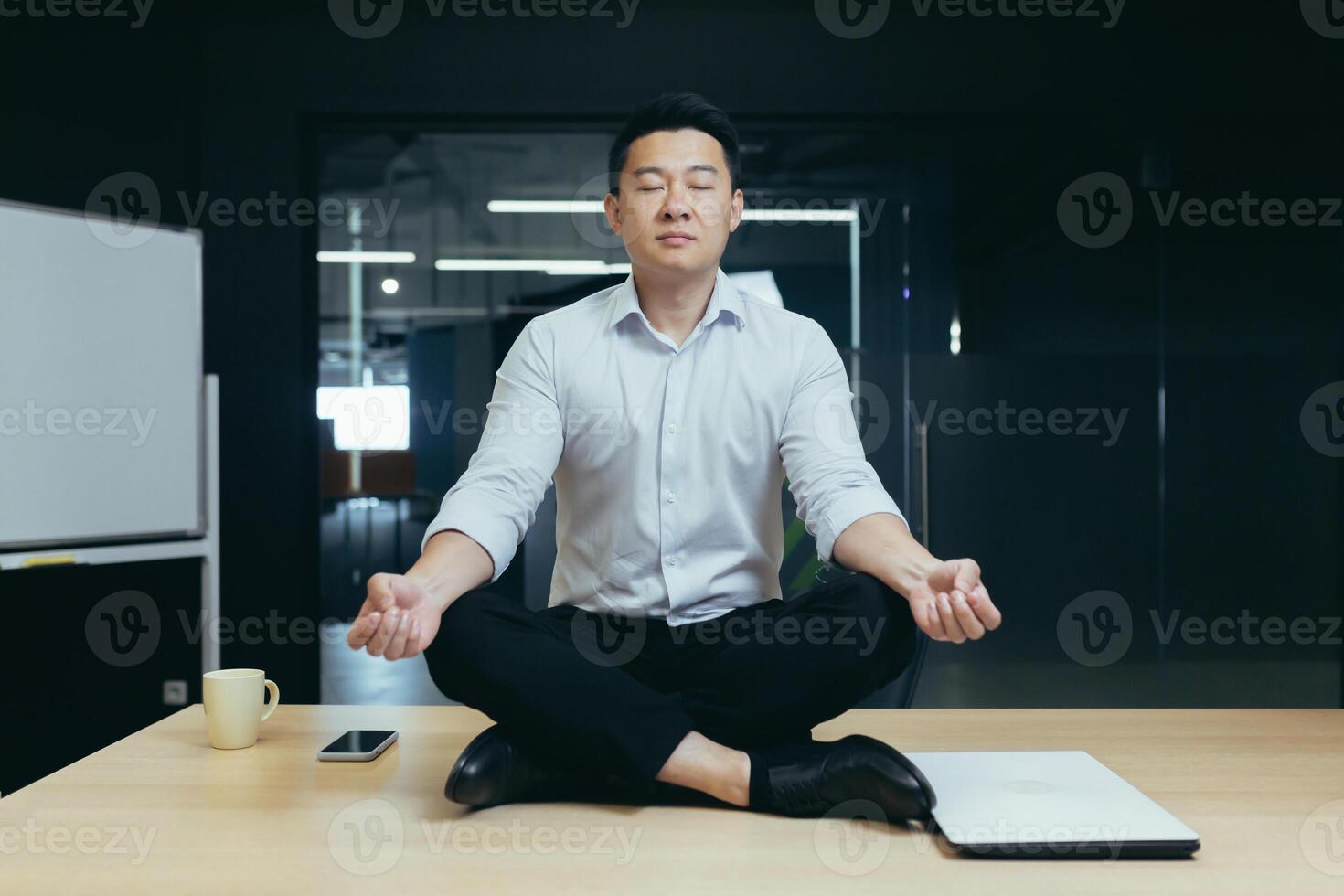 Rest break at the workplace. A young Asian man sits in the office on the desk in the lotus yoga position with closed eyes, meditates, relaxes during working hours. photo