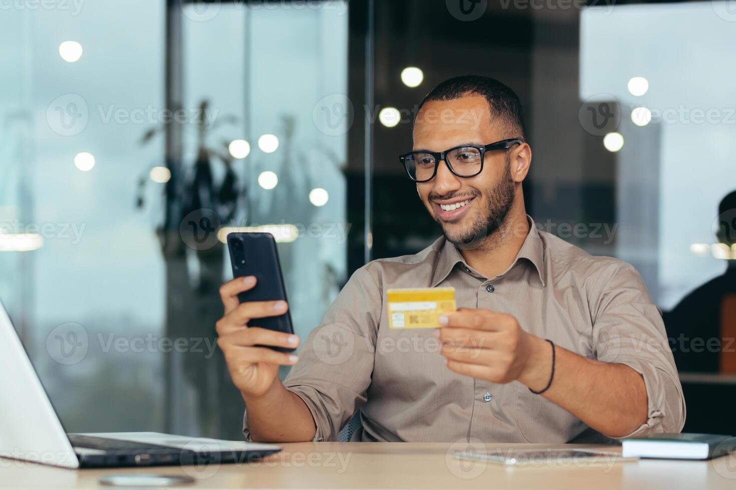 Happy african american man doing online shopping in online store remotely, businessman in glasses and shirt working inside modern office building with laptop, holding bank credit card and smartphone. photo