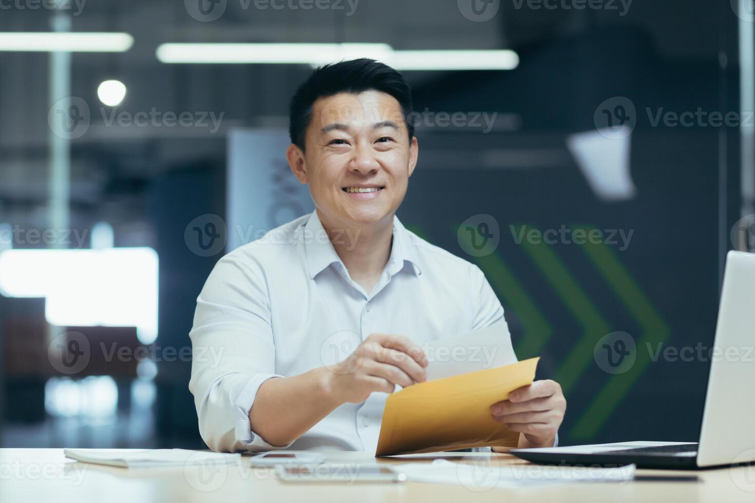 A young man, an Asian journalist, is sitting in the office at the table, holding an envelope with a letter, documents, an article in his hands. working on a laptop. He looks at the camera, smiles. photo