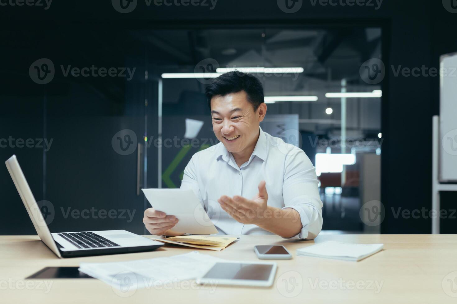 A young man, an Asian businessman, sits smiling in the office at the table, works with documents, holds papers, contracts, bills in his hands. photo
