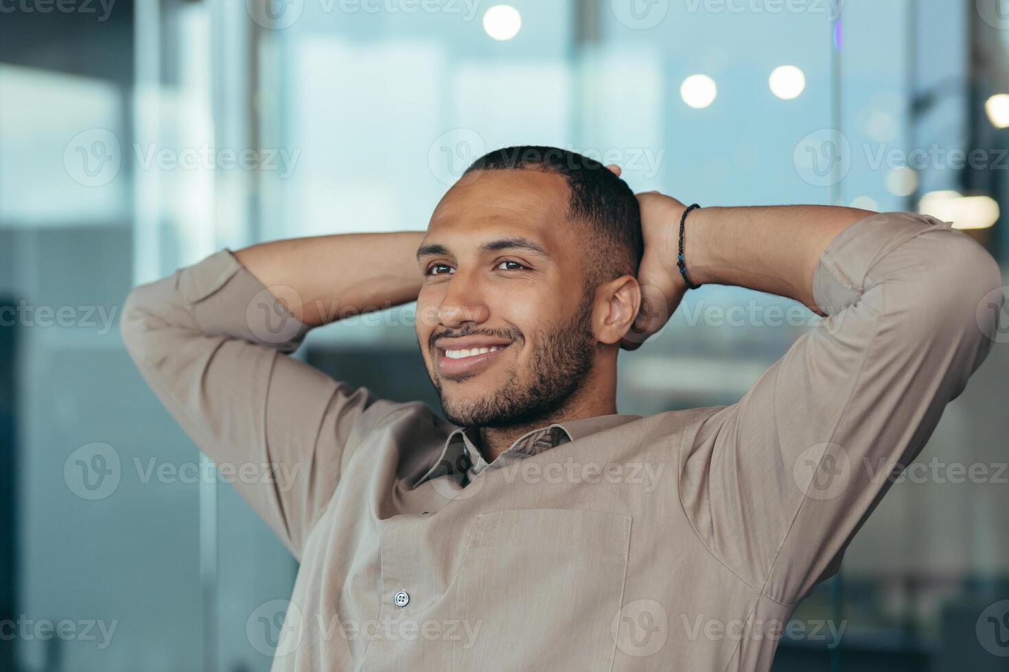 Close up photo of mixed race man relaxing in office with hands behind head looking out window and smiling, businessman daydreaming inside modern office building, successful investor happy with work